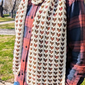 Può includere: Sciarpa all'uncinetto color crema con un motivo a cuore nei toni del marrone, arancione e rosso. La sciarpa è indossata sopra una camicia a quadri rossa e nera e jeans blu. Un accessorio accogliente.