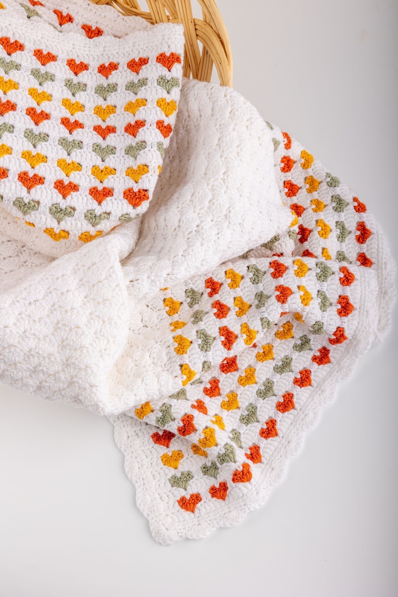 May include: A white crocheted blanket with a border of orange, yellow, and green hearts. The blanket is folded and draped over a wicker basket.