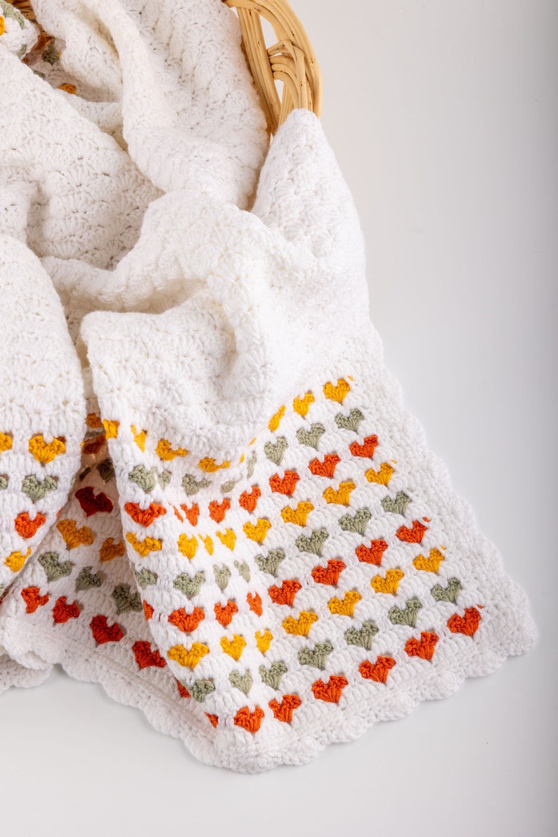 crochet baby heart blanket pattern with white yarn as base color in the middle with the shell stitch and autumn colors green orange and red for the hearts