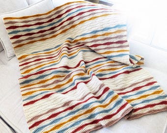 May include: A striped crocheted throw blanket in shades of beige, red, blue, and yellow. The blanket is folded and draped over a white couch.