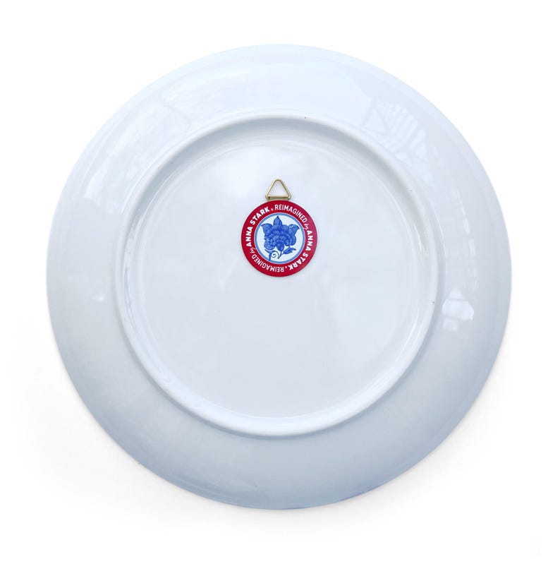 May include: White ceramic plate with a circular red and blue logo that reads "ANNA STARK REIMAGINED". The plate has a gold metal hanger attached to the back for display.