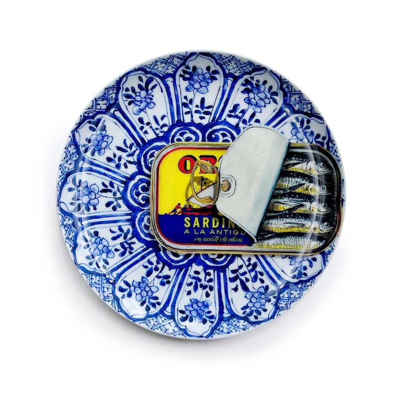 May include: A can of sardines, partially opened, rests on a blue and white patterned plate. The can has a yellow label with text and a picture of a boat. The plate has a floral design.
