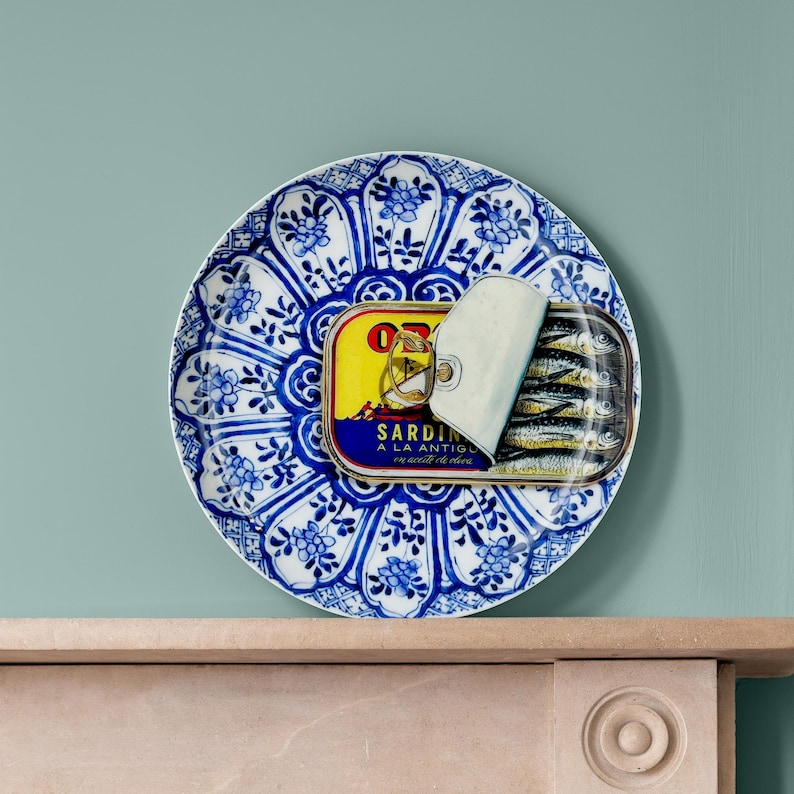 May include: A decorative blue and white plate with a floral design holds an open can of sardines. The sardine can has a yellow label with text and a silver lid. The sardines are visible inside the can.