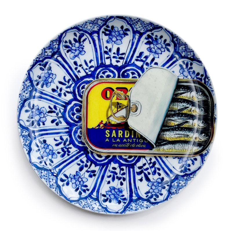 May include: A blue and white patterned ceramic plate with a can of sardines. The sardine can is yellow, red, and blue with the text "SARDIN A LA ANTIGU en aceite de oliva". The can is partially open, revealing the sardines.