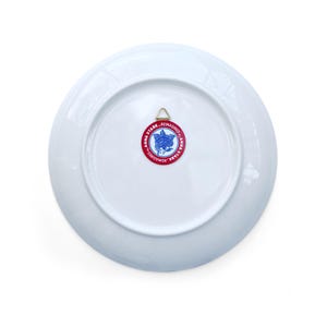 May include: A white ceramic plate with a gold hanging bracket on the back. A red and blue circular sticker with the text "ANNA STARK. REIMAGINED" surrounds a blue floral design.