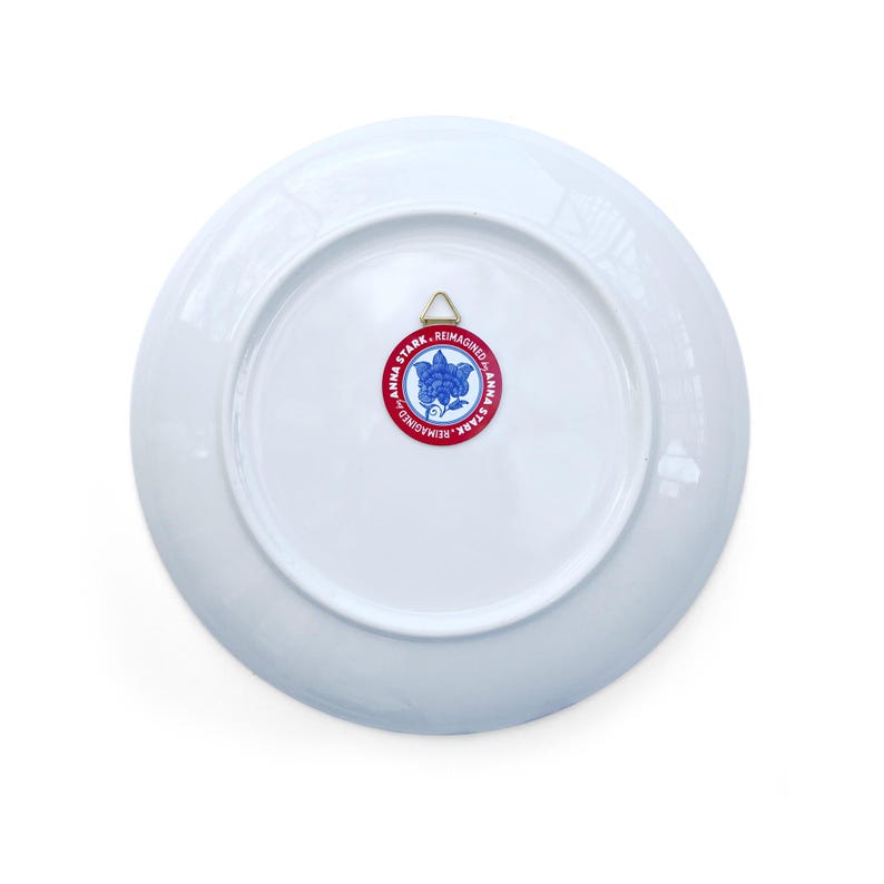 May include: A white ceramic plate with a gold hanging bracket on the back. A red and blue circular sticker with the text "ANNA STARK. REIMAGINED" surrounds a blue floral design.