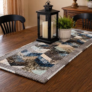 May include: A rectangular quilted table runner featuring a geometric pattern in shades of navy blue, light blue, cream, and charcoal gray. The runner has a solid gray border and is decorated with a repeating triangular design throughout the fabric.