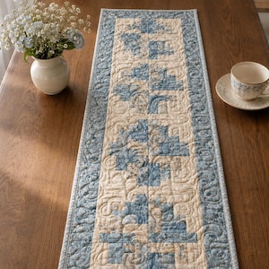 May include: A rectangular quilted table runner featuring a central panel of cream and blue geometric blocks surrounded by a border of blue fabric with intricate swirling stitch patterns. The runner has a light blue binding along the edges.
