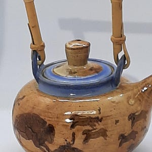 May include: A ceramic teapot with a brown and blue color scheme. The teapot features a bamboo handle and a lid with a small knob. The body of the teapot is decorated with brown animal illustrations.