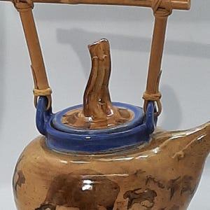 May include: A brown ceramic teapot with a blue lid and a bamboo handle. The teapot has a rustic, wood-like design and is decorated with a pattern of brown and white shapes.