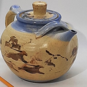 Può includere: Una teiera in ceramica con una glassa blu e un manico marrone. La teiera ha un coperchio marrone ed è decorata con un disegno marrone di animali che corrono.