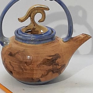 Op de afbeelding: Een bruine theepot van keramiek met een blauwe handgreep en deksel. De theepot heeft een gouden tuit en een uniek, abstract ontwerp.