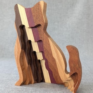 May include: A wooden cat sculpture with a layered design. The sculpture features various wood tones, including light brown, dark brown, purple, and cream. The cat is in a sitting position with its tail curled up. The sculpture is a decorative item.
