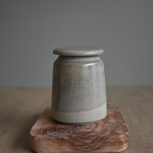Puede incluir: Un pequeño tarro de cerámica hecho a mano con tapa. El tarro es de color verde-beige apagado con una superficie ligeramente texturizada. Se encuentra sobre una tabla de cortar de madera con un patrón de vetas de madera natural. El tarro tiene forma cilíndrica.