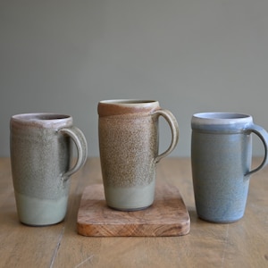 May include: Three handmade ceramic mugs in shades of green, brown, and blue. The mugs have a cylindrical shape with a handle and a textured surface. One mug sits on a small wooden board. The mugs are on a wooden table.