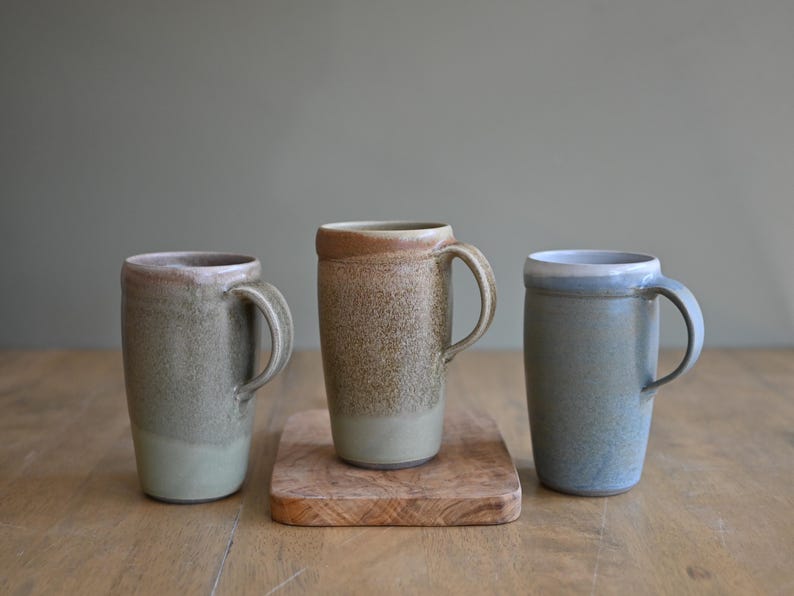 May include: Three handmade ceramic mugs in shades of green, brown, and blue. The mugs have a cylindrical shape with a handle and a textured surface. One mug sits on a small wooden board. The mugs are on a wooden table.