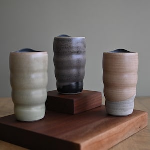 May include: Three ceramic tumblers with a wavy design in shades of beige, gray, and black. The tumblers are displayed on a dark wooden stand. The background is a muted green.