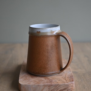 May include: A brown ceramic mug with a white rim and a handle. The mug is sitting on a wooden surface.