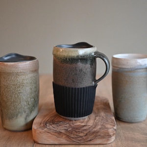 May include: Three ceramic mugs in earthy tones sit on a wooden surface. The mugs vary in shape and color, with one featuring a black silicone sleeve. The mugs have handles and are suitable for hot beverages.