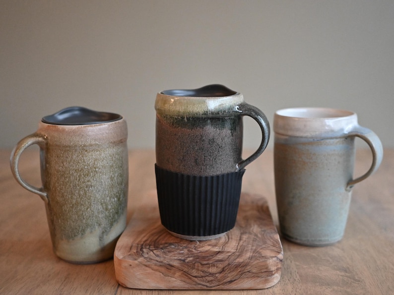 May include: Three ceramic mugs in earthy tones sit on a wooden surface. The mugs vary in shape and color, with one featuring a black silicone sleeve. The mugs have handles and are suitable for hot beverages.