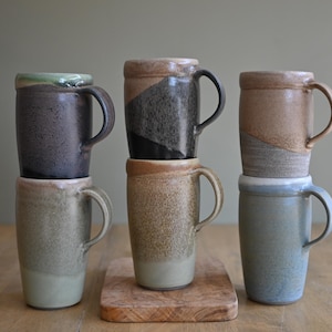 May include: Six ceramic mugs in various colors, including gray, brown, and blue, are stacked on a wooden surface. Each mug has a handle and a unique glaze, with some featuring color gradients and textured finishes. The mugs are approximately 5 inches tall.