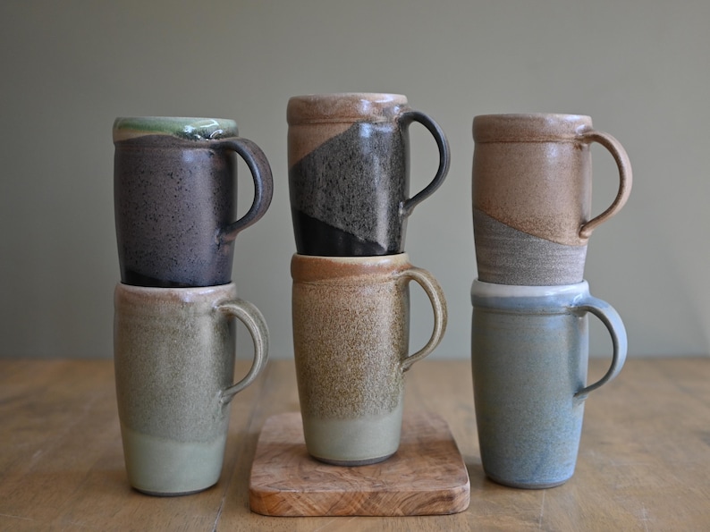 May include: Six ceramic mugs in various colors, including gray, brown, and blue, are stacked on a wooden surface. Each mug has a handle and a unique glaze, with some featuring color gradients and textured finishes. The mugs are approximately 5 inches tall.