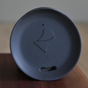 May include: A dark gray lid with a curved design and the word "Portava" printed on it. The lid has a small hole and a curved indentation. The lid is on a wooden surface.