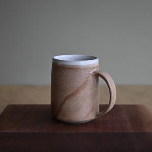 May include: A cylindrical ceramic mug with a speckled tan and brown exterior finish. The mug features a smooth white glazed rim and a matching curved handle on the right side. The body of the mug displays a subtle, swirling brown pattern.