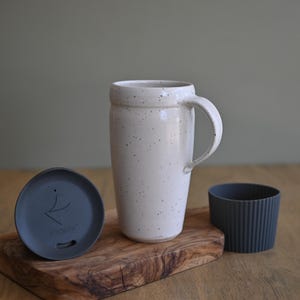 May include: A cream-colored ceramic travel mug with a handle, speckled with dark spots, sits on a wooden board. A dark gray lid and a ribbed silicone sleeve are also present. The mug is designed for hot or cold beverages.