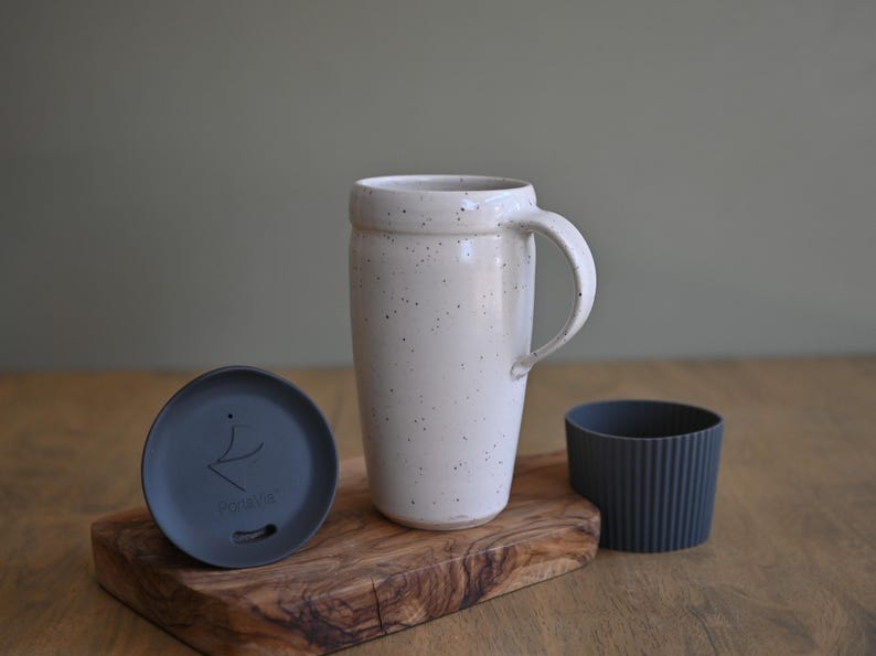 May include: A cream-colored ceramic travel mug with a handle, speckled with dark spots, sits on a wooden board. A dark gray lid and a ribbed silicone sleeve are also present. The mug is designed for hot or cold beverages.