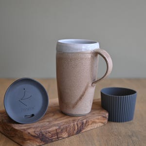May include: A ceramic travel mug with a handle, a lid, and a silicone sleeve. The mug is tan with a white rim. The lid is dark gray with the text "Portavita". The sleeve is dark gray and ribbed. The items are on a wooden cutting board.