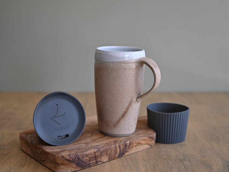 May include: A ceramic travel mug with a handle, a lid, and a silicone sleeve. The mug is tan with a white rim. The lid is dark gray with the text "Portavita". The sleeve is dark gray and ribbed. The items are on a wooden cutting board.
