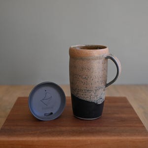 May include: A tall ceramic travel mug with a speckled tan and brown upper section and a solid black base, featuring a curved handle. Beside the mug sits a circular dark gray silicone lid with a small drinking slot and the text Portavia printed on it.