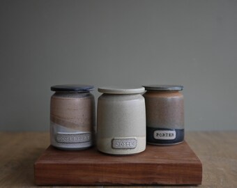 May include: Three cylindrical ceramic jars with lids, each featuring a rectangular label. The left jar is tan and brown with the text GOOSEBERRY, the center jar is cream with the text JOSTE, and the right jar is brown and black with the text PORTER.