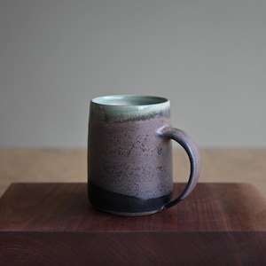 Könnte beinhalten: Handgefertigte Keramiktasse mit dunkelbrauner und schwarzer gesprenkelter Glasur. Der Innenrand ist hellgrün. Die Tasse hat einen gebogenen Henkel und steht auf einer dunkelbraunen Holzoberfläche.