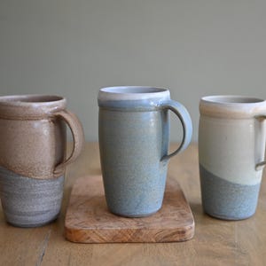May include: Three handmade ceramic mugs in neutral tones. The mugs have a cylindrical shape with a curved handle. The colors include beige, gray, and blue, with a two-tone design. One mug sits on a small wooden cutting board.