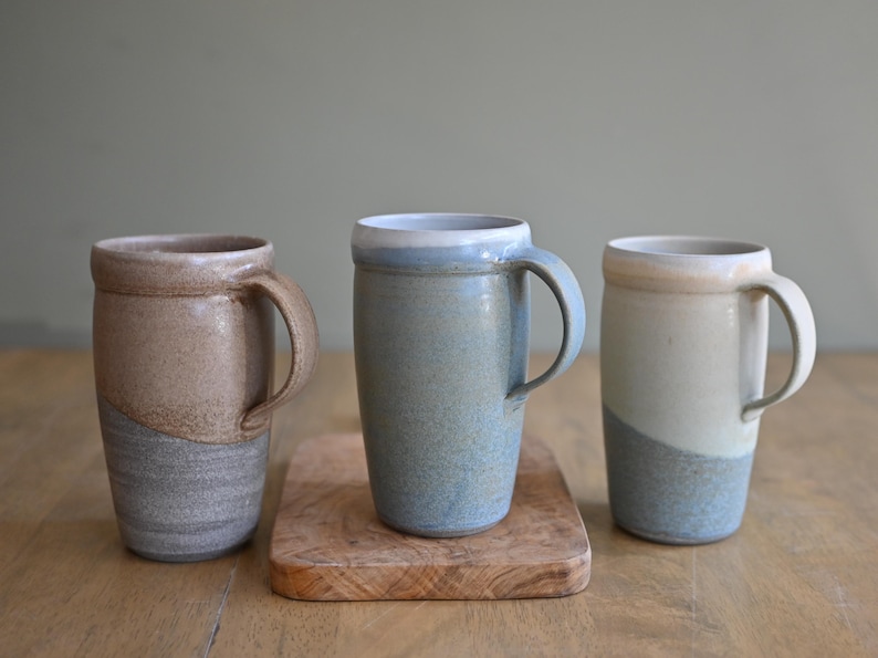 May include: Three handmade ceramic mugs in neutral tones. The mugs have a cylindrical shape with a curved handle. The colors include beige, gray, and blue, with a two-tone design. One mug sits on a small wooden cutting board.
