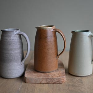 Puede incluir: Tres jarras de cerámica en diferentes colores y esmaltes. Las jarras están sobre una superficie de madera. Las jarras son todas del mismo tamaño y forma, pero tienen diferentes colores y esmaltes. La jarra de la izquierda es de color gris claro con un esmalte moteado. La jarra del centro es de color marrón con un esmalte moteado. La jarra de la derecha es de color verde claro con un esmalte liso.