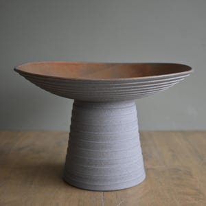 May include: A grey ceramic pedestal bowl with a wide, shallow dish and a textured, ribbed design. The interior of the bowl is a warm, earthy brown. The bowl is elevated on a cylindrical base with horizontal grooves, creating a modern, minimalist aesthetic.