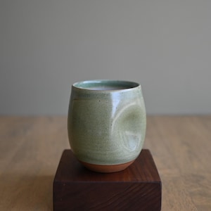 Puede incluir: Un vaso de cerámica hecho a mano con forma redondeada y un esmalte verde moteado. La base es de color marrón cálido. La taza está sobre un bloque de madera oscura, con un fondo neutro.