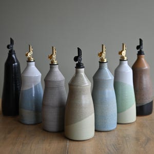Peut inclure: Une collection de sept distributeurs d'huile en céramique disposés en demi-cercle. Chaque bouteille présente une couleur d'émail unique, notamment noir, blanc, bleu, gris et beige, avec un bec verseur en métal doré et un bouchon noir.