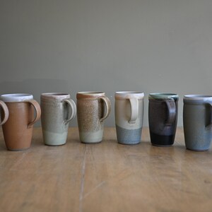 May include: A collection of seven handmade ceramic mugs with unique color variations. The mugs have a cylindrical shape with a handle and are displayed on a wooden surface. The colors range from light beige to dark blue and black.
