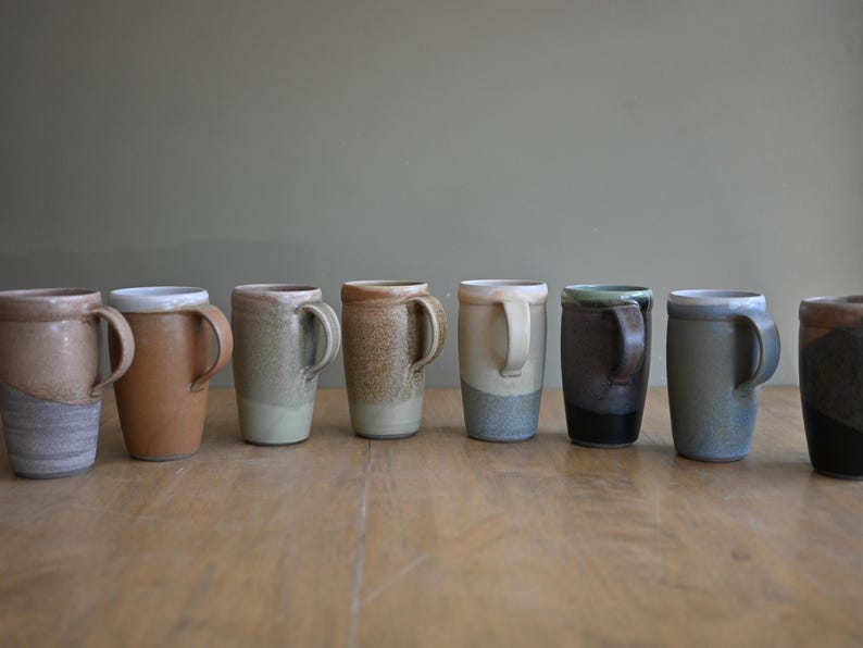 May include: A collection of seven handmade ceramic mugs with unique color variations. The mugs have a cylindrical shape with a handle and are displayed on a wooden surface. The colors range from light beige to dark blue and black.