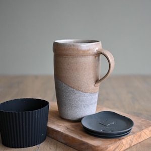 May include: A two-tone ceramic travel mug with a handle, in beige and grey. A black silicone sleeve and a dark grey lid sit beside the mug on a wooden board. The mug is designed for hot or cold drinks, and is suitable for travel.