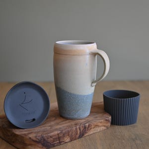 May include: A handmade ceramic travel mug with a handle, featuring a cream, tan, and blue color scheme. Includes a matching lid and a dark gray ribbed sleeve. The lid has the text "PortaVie". The mug sits on a wooden cutting board.