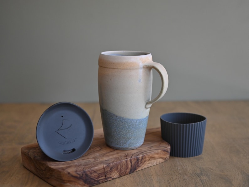 May include: A handmade ceramic travel mug with a handle, featuring a cream, tan, and blue color scheme. Includes a matching lid and a dark gray ribbed sleeve. The lid has the text "PortaVie". The mug sits on a wooden cutting board.