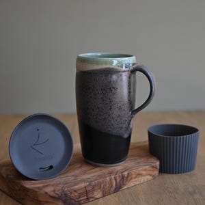 May include: A tall ceramic mug with a black handle, a dark gray speckled body, and a light green rim. Includes a dark gray lid and a ribbed gray silicone sleeve. The mug sits on a wooden cutting board.