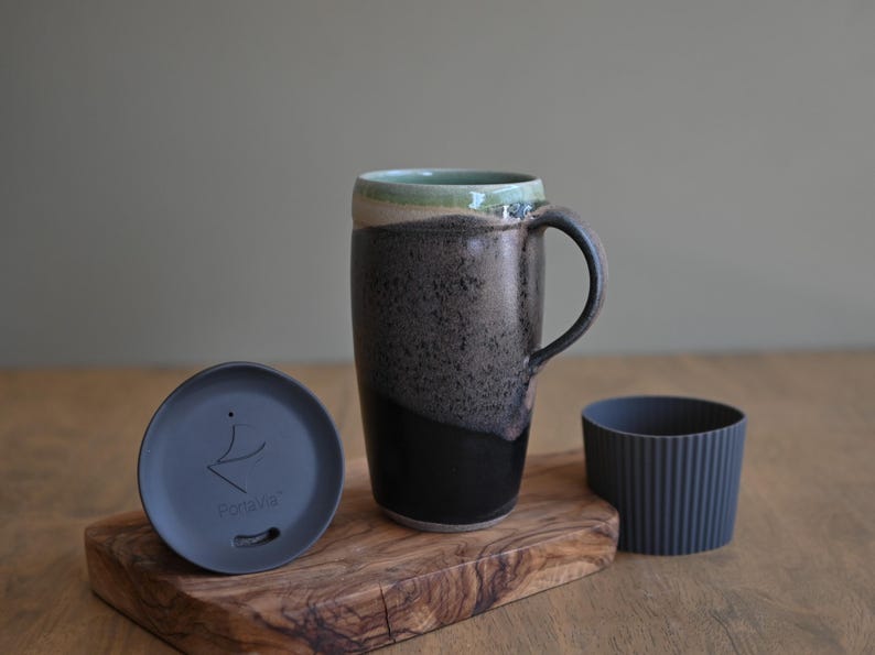 May include: A tall ceramic mug with a black handle, a dark gray speckled body, and a light green rim. Includes a dark gray lid and a ribbed gray silicone sleeve. The mug sits on a wooden cutting board.