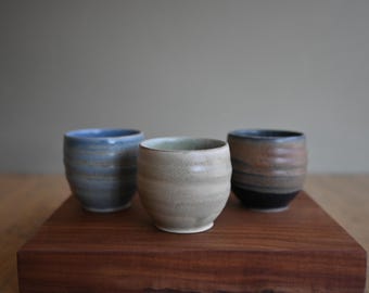 May include: Three handcrafted ceramic cups in shades of blue, beige, and brown, each with a unique textured surface. The cups are displayed on a dark wooden tray, showcasing their artisanal quality and earthy tones.