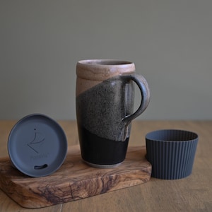 May include: A ceramic travel mug with a handle, a lid, and a silicone sleeve. The mug is two-toned, with a black base and a gray and tan upper section. The lid is dark gray and has a logo. The sleeve is dark gray.